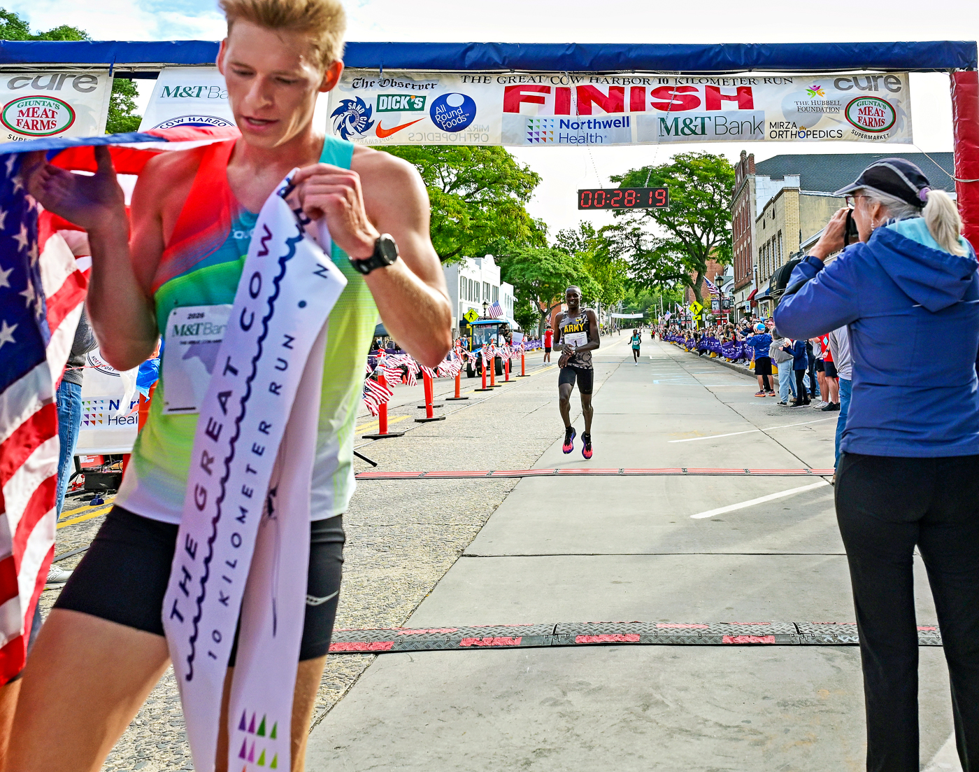1st-Place Winner Chandler Gibbens With The Finish Tape and 2nd-Place Winner Anthony Rotich Crossing The Finish Line. The Great Cow Harbor 10K Run 2026